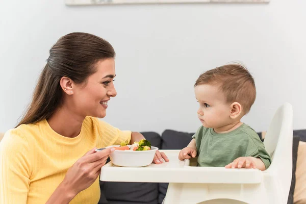 Vista lateral de la madre sonriente sosteniendo un tazón de verduras cerca de su hijo pequeño en la silla alta - foto de stock