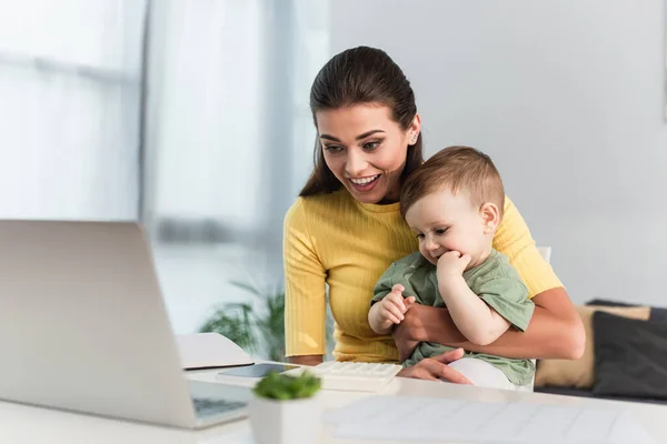 Mujer alegre sosteniendo hijo pequeño cerca de la computadora portátil en primer plano borrosa - foto de stock