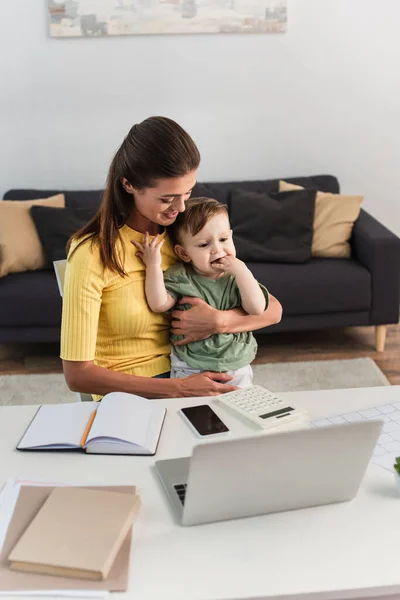 Sonriente freelancer abrazando hijo cerca de dispositivos y cuadernos en casa - foto de stock