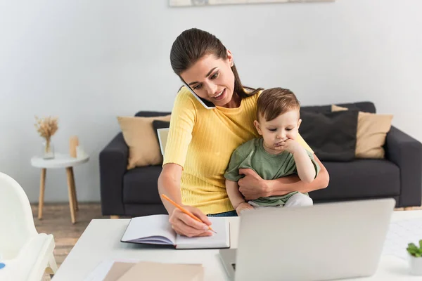 Freelancer talking on smartphone and writing on notebook near laptop and son — Stock Photo