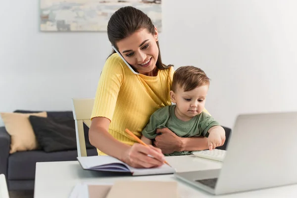 Smiling mother writing on notebook and talking on smartphone near laptop and toddler son — Stock Photo