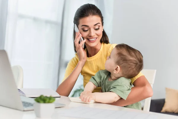 Smiling mother talking on smartphone near toddler son with calculator — Stock Photo