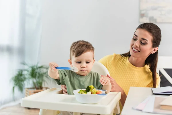 Lächelnde Mutter schaut Sohn mit Löffel in Gemüseschüssel auf Hochstuhl an — Stockfoto