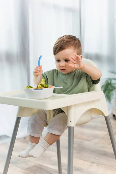 Kleinkind hält Löffel in der Nähe verschwommene Schüssel mit Gemüse auf Hochstuhl — Stockfoto