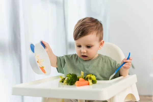Criança segurando colher e tigela perto de legumes na cadeira alta — Fotografia de Stock