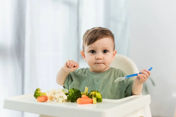 Bambino ragazzo tenendo cucchiaio e guardando la fotocamera vicino verdure sul seggiolone — Foto stock