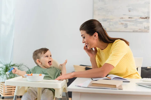 Femme souriante parlant sur un téléphone mobile près du fils qui sort la langue et de la nourriture sur une chaise haute — Photo de stock