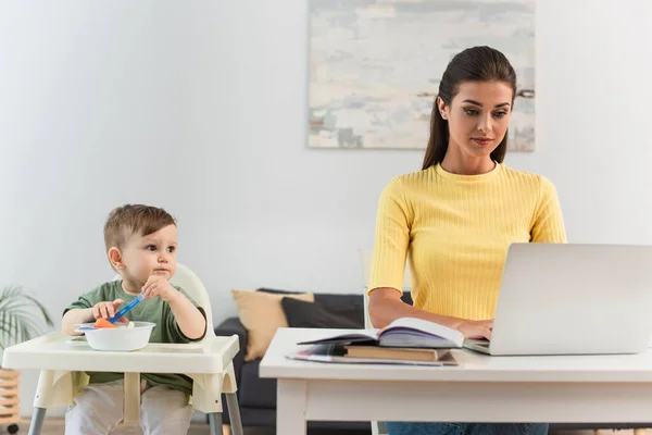 Freelance utilisant un ordinateur portable près de son fils avec de la nourriture sur chaise haute à la maison — Photo de stock