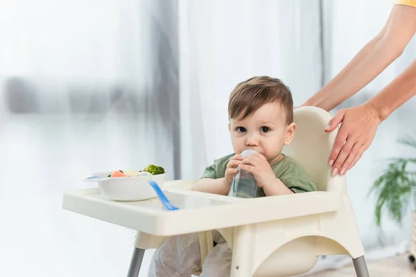 Mulher de pé perto do filho da criança com mamadeira e comida na cadeira alta — Fotografia de Stock