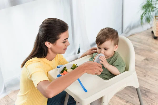 Madre sosteniendo biberón con chupete cerca del niño y tazón de verduras en la silla alta - foto de stock