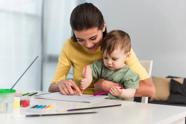 Jeune femme souriant tout en pointant vers le papier près de son fils avec des crayons à la maison — Photo de stock