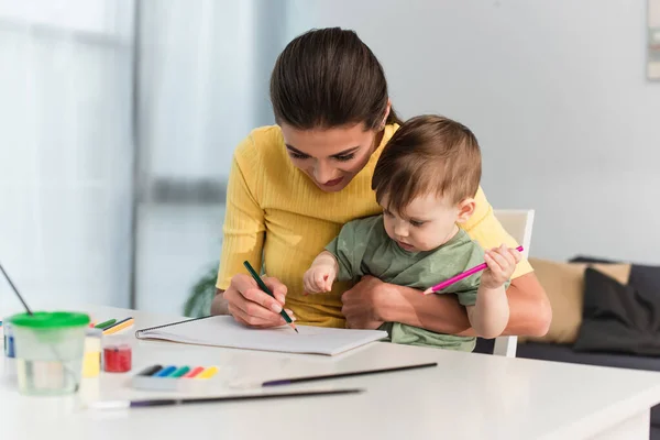 Woman drawing with toddler kid near gouache on blurred foreground — Stock Photo