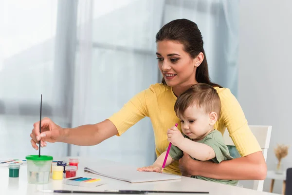 Femme souriante tenant un pinceau près de l'enfant avec un crayon — Photo de stock