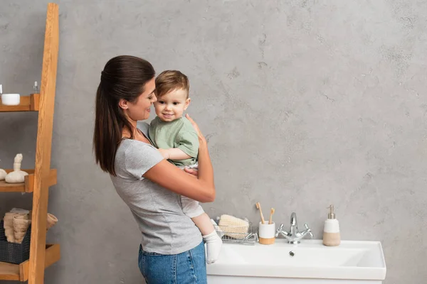 Alegre madre abrazando hijo pequeño cerca del lavabo en el baño - foto de stock