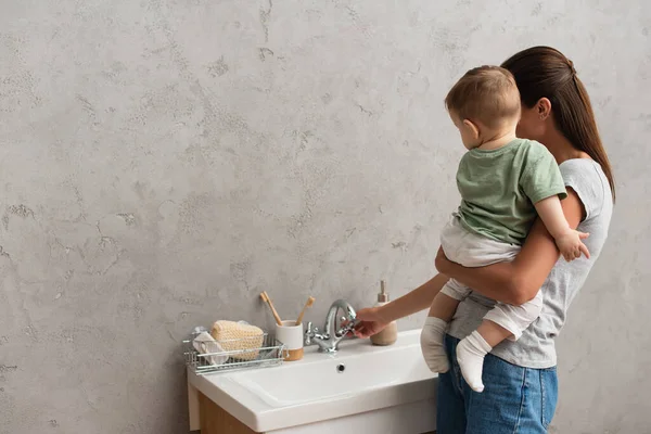 Mother opening tap of faucet while holding toddler kid in bathroom — Stock Photo