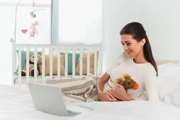 Smiling woman holding toy near laptop on bed — Stock Photo