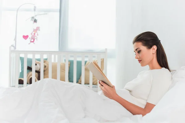 Seitenansicht einer Frau, die Buch auf dem Bett in der Nähe von verschwommenem Babybett liest — Stockfoto