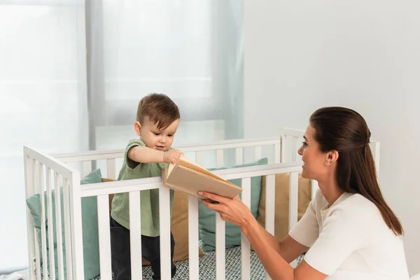 Mujer sonriente sosteniendo libro cerca de niño en cama de bebé — Stock Photo