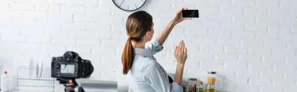 Домогосподарка махає рукою біля смартфона з порожнім екраном і розмитою цифровою камерою на кухні, банер — стокове фото