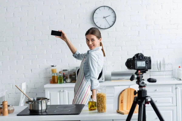 Усміхнена домогосподарка бере селфі біля плити і розмиту цифрову камеру на кухні — стокове фото