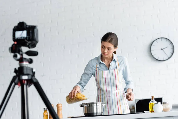 Blogueiro culinário em avental preparando macarrão na frente de câmera digital borrada — Fotografia de Stock