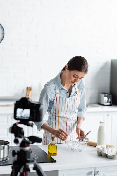 Pretty culinary blogger breaking chicken egg into bowl in front of blurred digital camera — Stock Photo