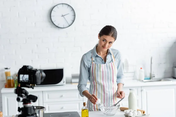 Pretty housewife looking at digital camera while breaking chicken egg into bowl — Stock Photo