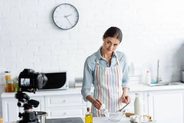 Hübsche Hausfrau schaut auf Digitalkamera, während sie Hühnerei in Schüssel bricht — Stockfoto