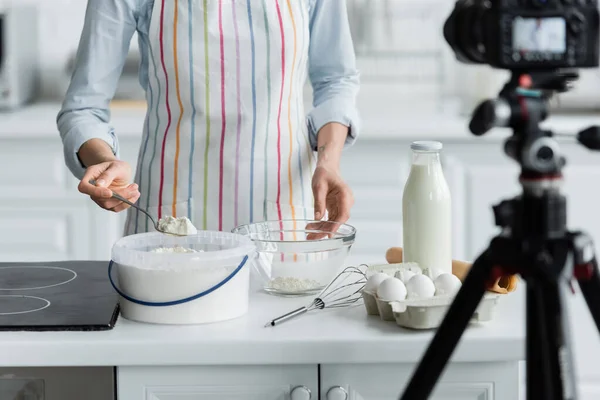 Abgeschnittene Ansicht einer Frau in Schürze, die Mehl in eine Schüssel in der Nähe einer verschwommenen Digitalkamera gibt — Stockfoto