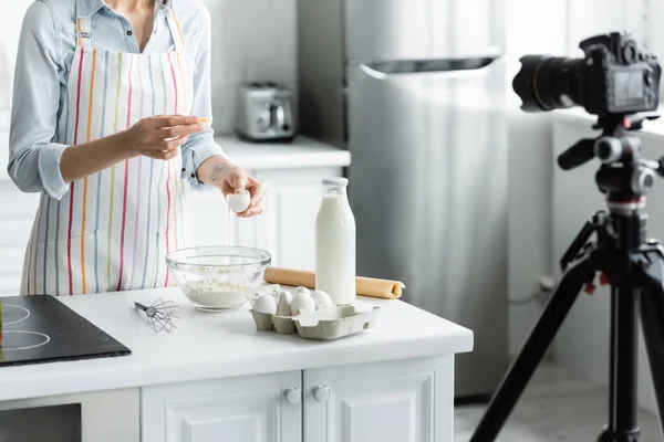 Cropped view of woman breaking egg into bowl with flour near blurred digital camera — Stock Photo