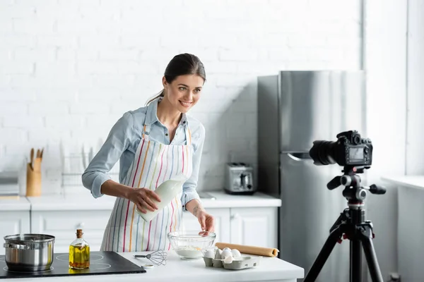 Smiling housewife holding bottle of milk near bowl with flour in front of digital camera — Stock Photo