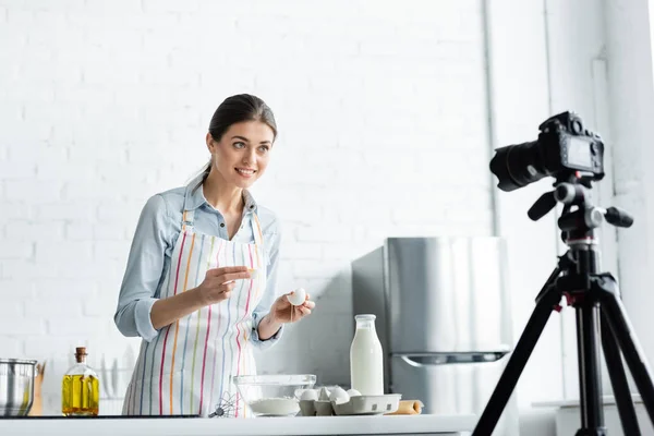 Happy culinary blogger looking at blurred digital camera while breaking chicken egg into bowl with flour — Stock Photo