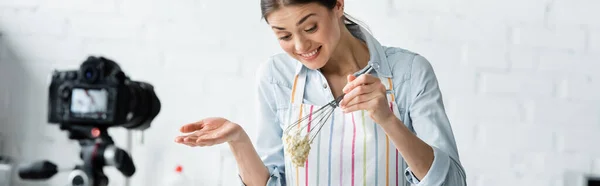 Cheerful culinary blogger holding whisk with raw dough near blurred digital camera, banner — Stock Photo