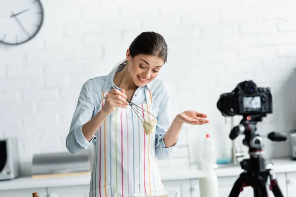 Joyeuse ménagère tenant fouet avec de la pâte pendant la cuisson en ligne près de l'appareil photo numérique flou — Photo de stock