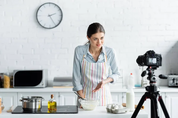 Joven ama de casa apuntando a cuenco con harina cerca de cámara digital durante la cocina en línea - foto de stock