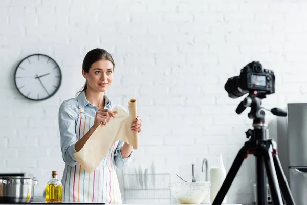 Blogger culinario en delantal sosteniendo pergamino para hornear cerca de un tazón con masa y cámara digital borrosa - foto de stock