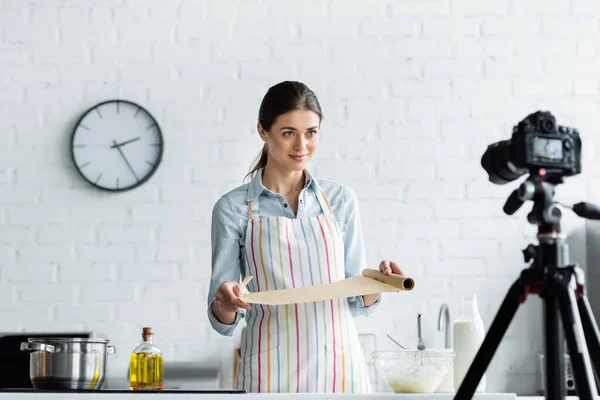 Blogger culinario sosteniendo pergamino para hornear cerca de un tazón con masa y cámara digital borrosa - foto de stock