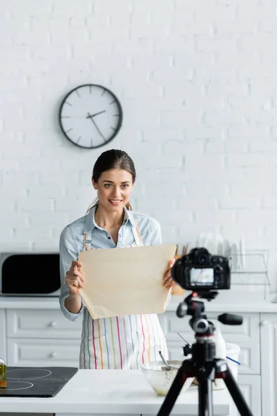 Blogueur culinaire souriant montrant le parchemin de cuisson à l'appareil photo numérique flou dans la cuisine — Photo de stock