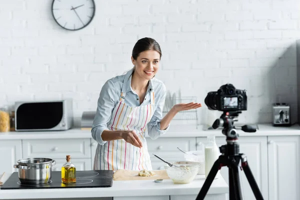 Cheerful culinary blogger pointing with hand near dough and blurred digital camera — Stock Photo