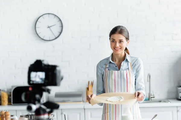 Fröhliche kulinarische Bloggerin zeigt Backpapier mit rohem Teig in der Nähe einer unscharfen Digitalkamera — Stockfoto