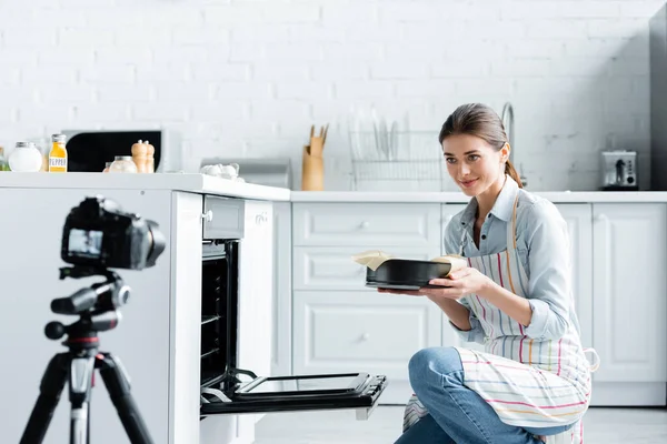 Blurred digital camera near young culinary blogger holding baking form near oven — Stock Photo