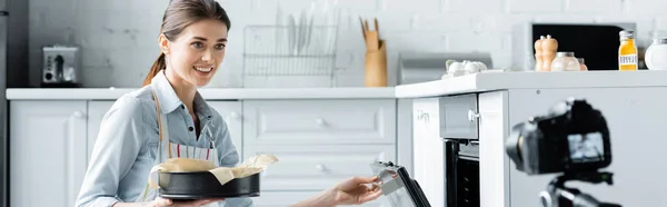 Sonriente blogger culinario abriendo el horno mientras mantiene la forma de hornear cerca de la cámara digital borrosa, pancarta - foto de stock