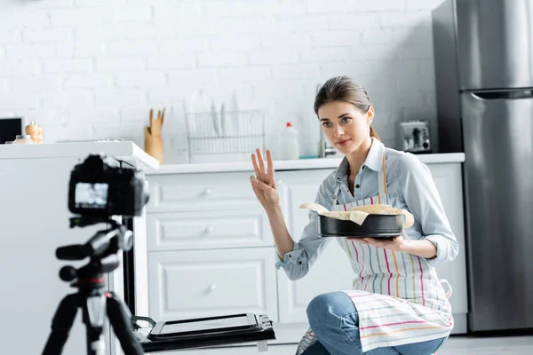 Culinary blogger showing cooking time while holding baking form near blurred digital camera — Stock Photo
