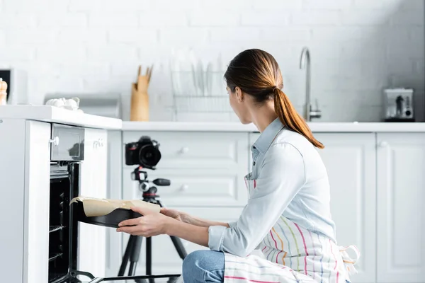Junge Frau legt Backform in Backofen, während sie in der Küche auf Digitalkamera blickt — Stockfoto