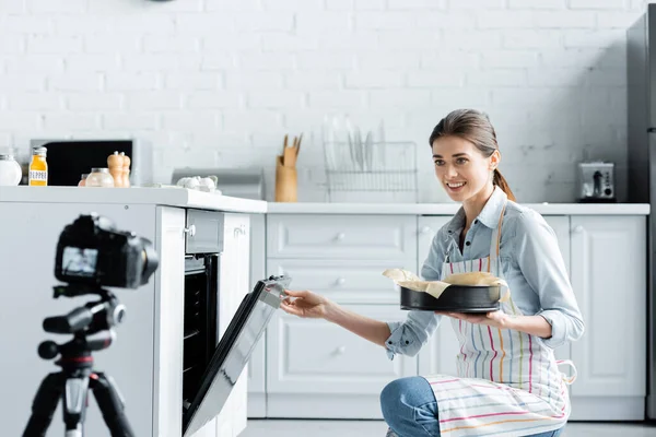 Smiling culinary blogger holding baking form near oven and blurred digital camera — Stock Photo