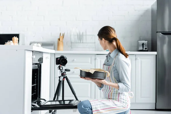 Jeune femme au foyer tenant forme de cuisson près du four et appareil photo numérique dans la cuisine — Photo de stock