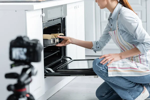 Ausgeschnittene Ansicht einer kulinarischen Bloggerin, die Backform in den Ofen legt, in der Nähe einer verschwommenen Digitalkamera — Stockfoto