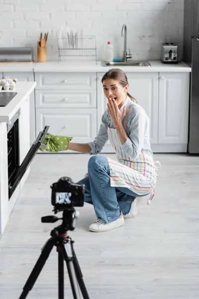 Изумленный кулинарный блогер, прикрывающий рот, открывая духовку рядом с размытой цифровой камерой — стоковое фото