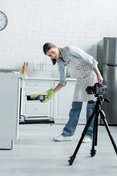 Visão de comprimento total da mulher no avental colocando forma de cozimento no forno perto da câmera digital no tripé — Fotografia de Stock