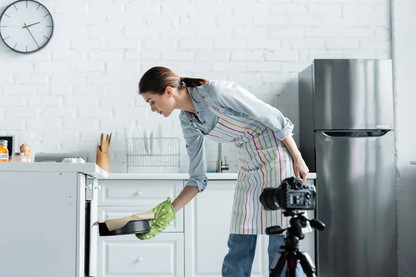 Giovane casalinga mettendo la forma di cottura in forno vicino alla fotocamera digitale offuscata — Foto stock